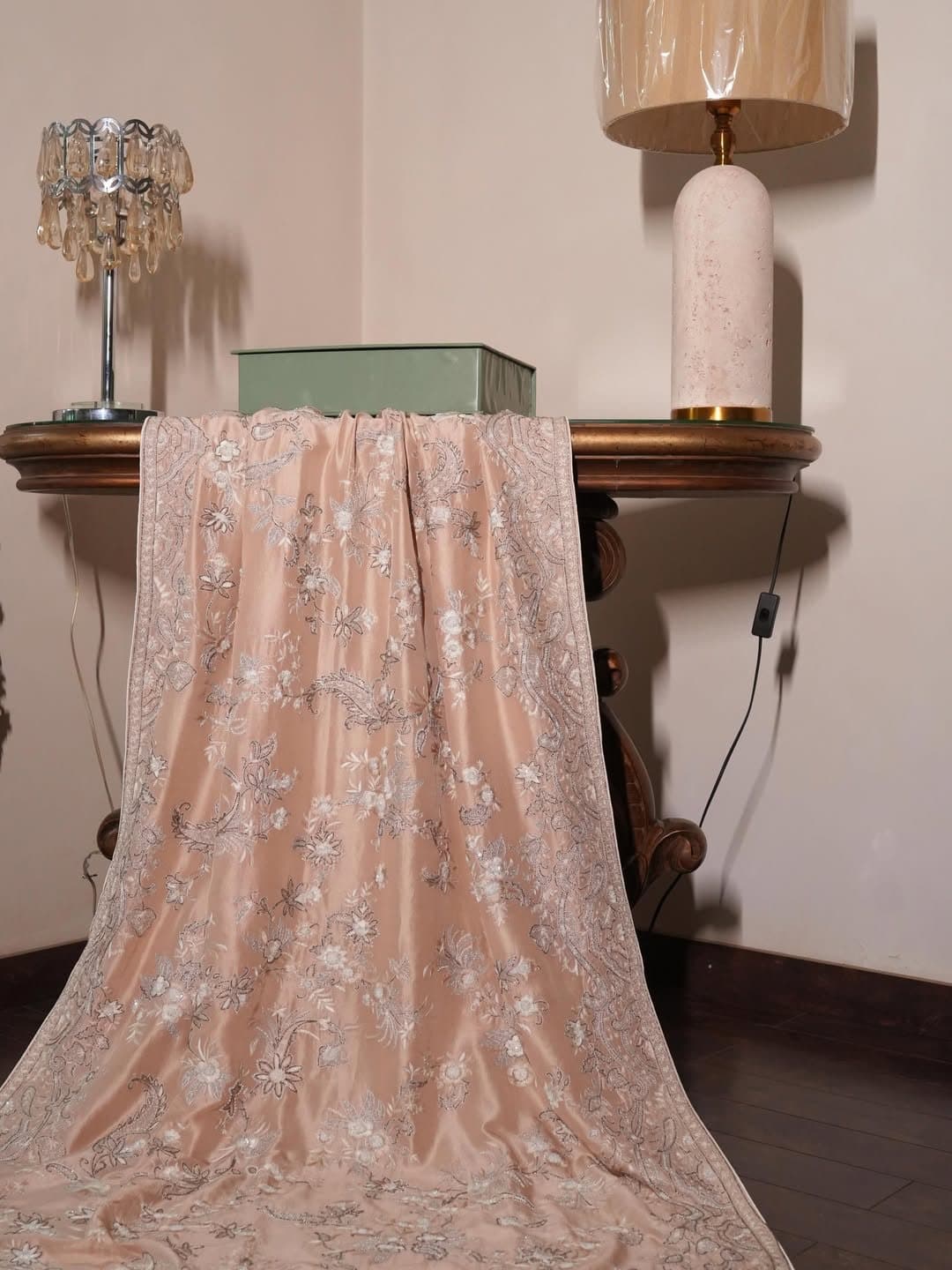 A blush shawl with fine pale embroidery, draped over a console in a softly lit room.