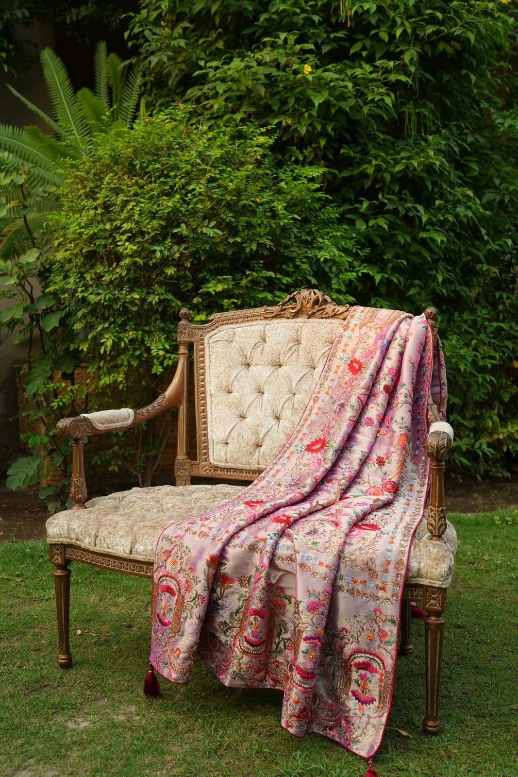 A rose kalamkaar shawl with multicoloured floral embroidery, draped over a carved antique chair in a garden.