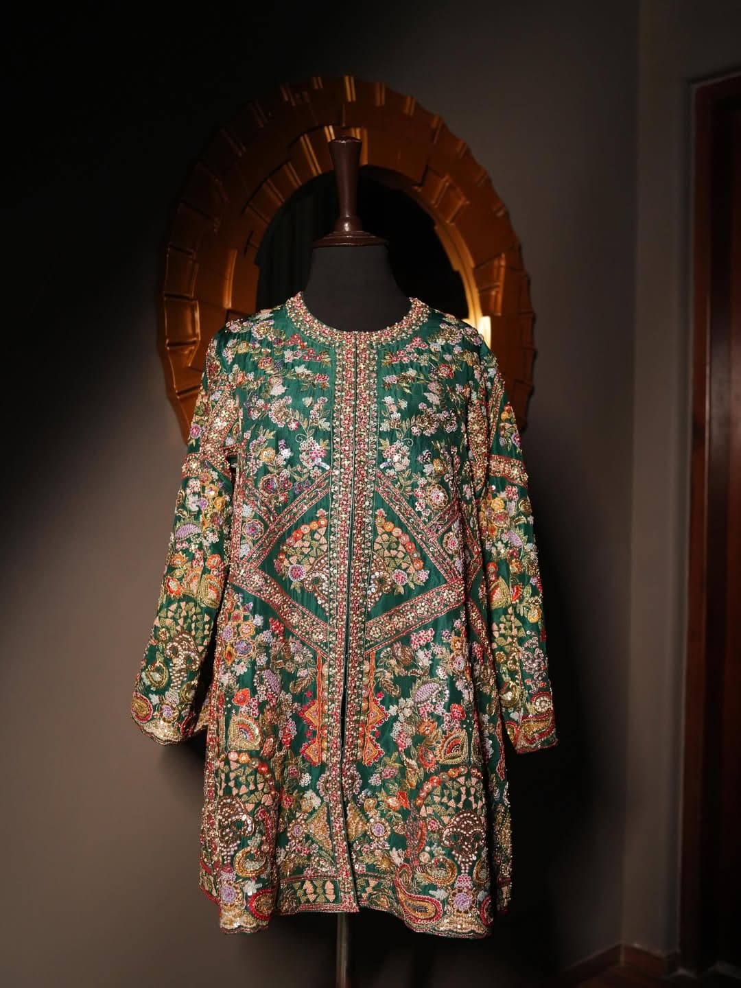 A darkly lit embroidered coat on a stand, its surface worked in metallic thread.