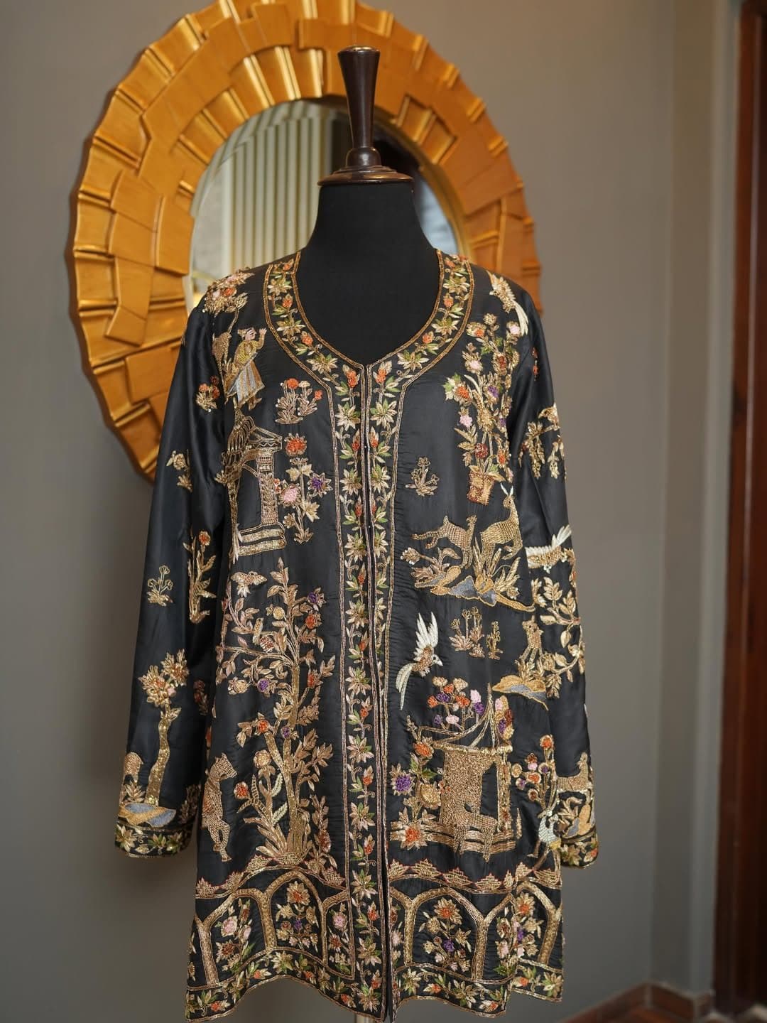 A dark jacket with gold and jewel-toned embroidery, displayed against a gilded arch.